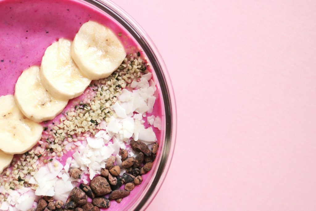 A brightly colored smoothie bowl with fresh banana, seeds, and coconut flakes on a pink background.